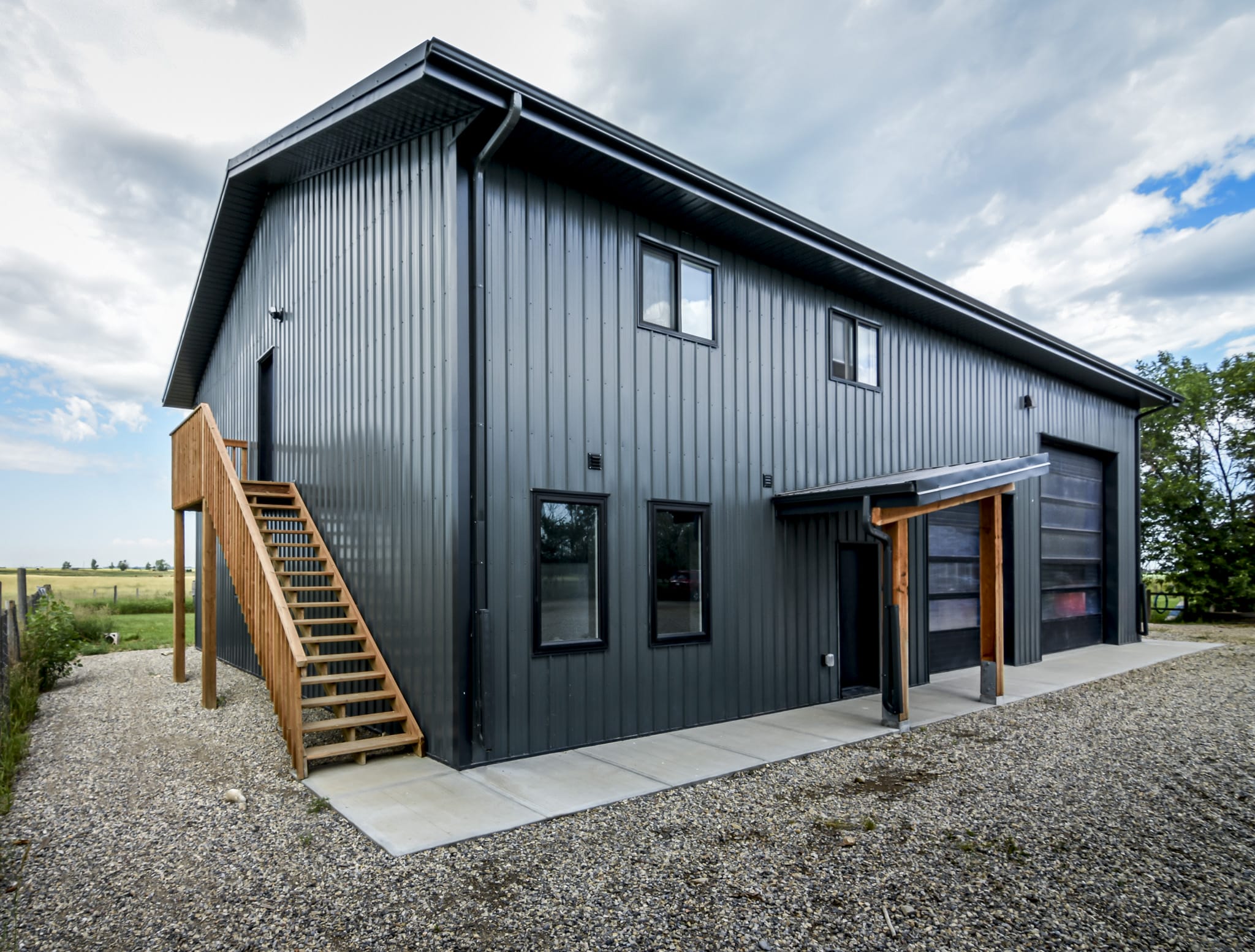 Modern multi-purpose post frame acreage building with garage, flight of stairs leading up to a second storey side entrance and black metal exterior