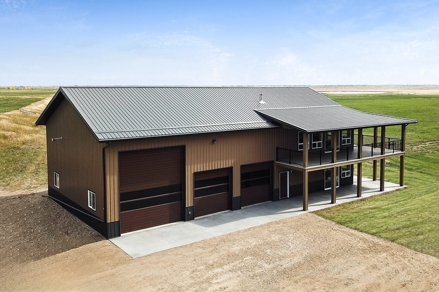 Modern barndominium with three overhead doors, a veranda and brown exterior.