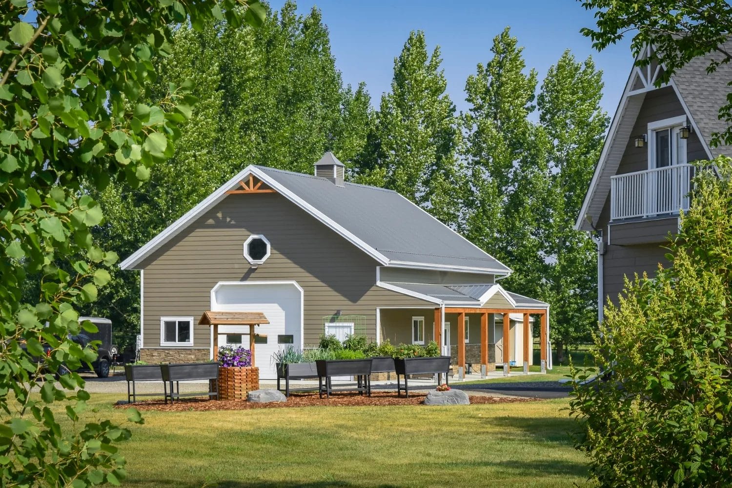 A premium post frame garage sits on a treed acreage lot. The building was design with upgraded features to match the aesthetic of the main home, which is seen at the right edge of the photo.
