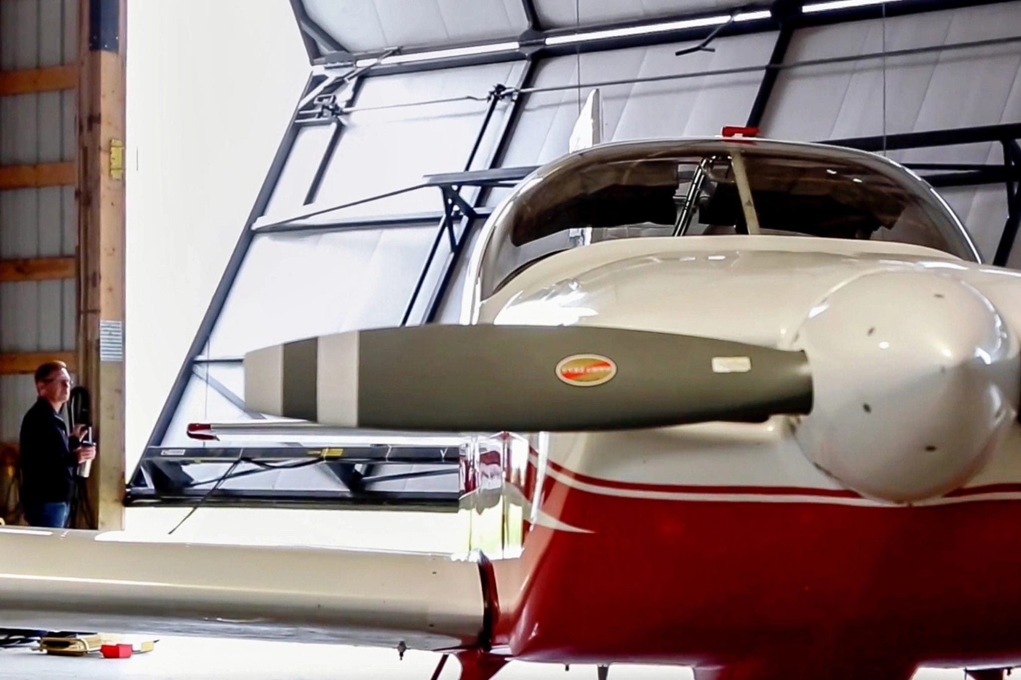 A small aircraft pilot opening the bi-fold door of a post frame airplane hangar in the background. A small aircraft sits in the foreground, ready to fly.