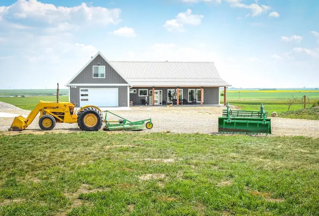 Small tractors and equipment sit in front of a 4 bedroom barndominium with loft above the garage